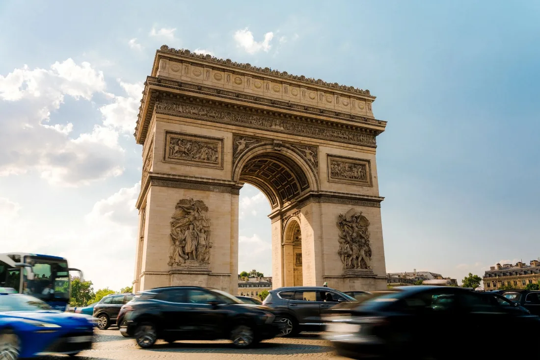 Triumphal stone arch under bright cloud with cars and a bus streaking past in motion blur across the foreground.