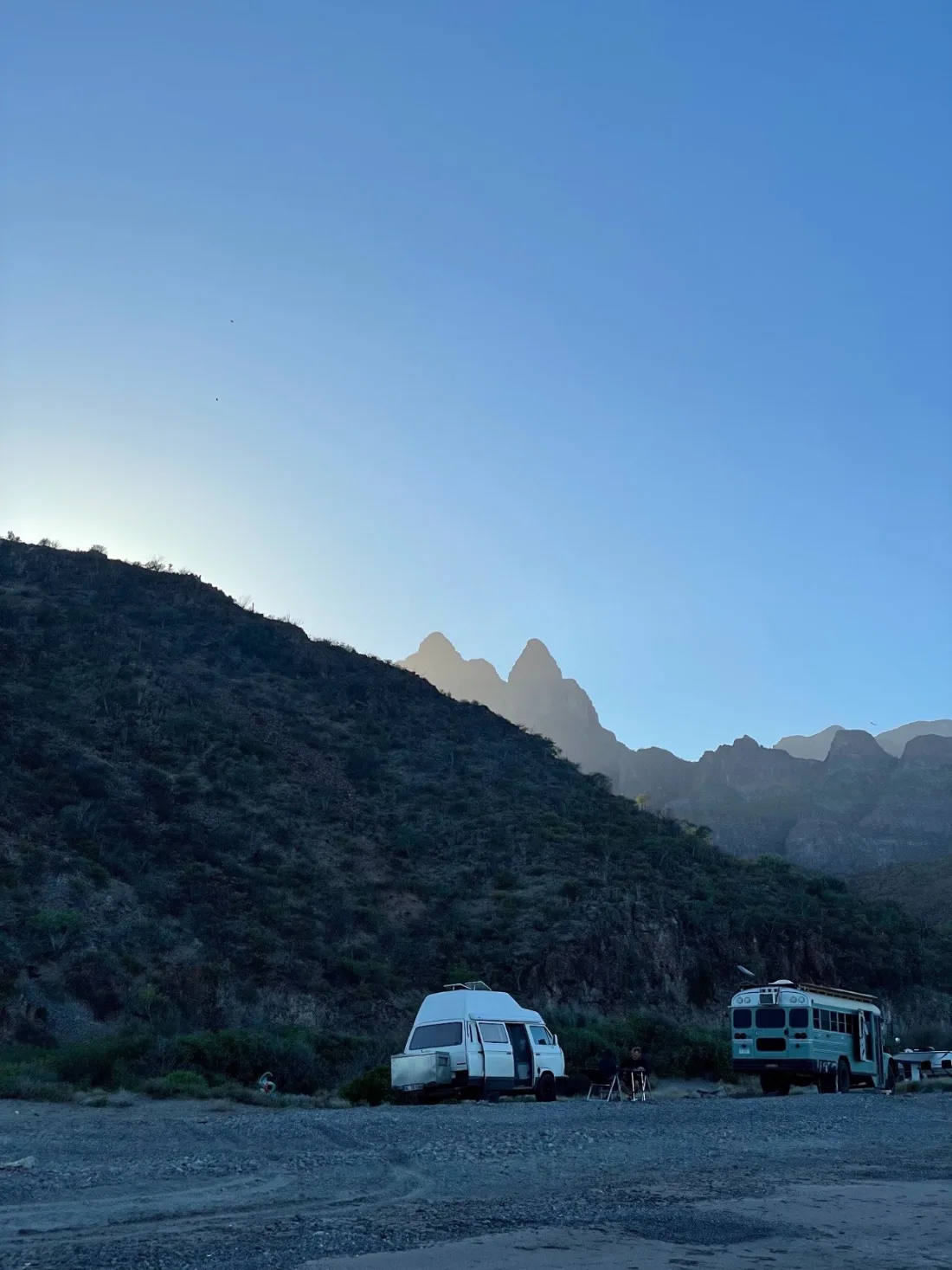 A camper van and a converted school bus parked on a gravel beach at blue hour, cactus-covered ridges and hazy spires rising behind them.
