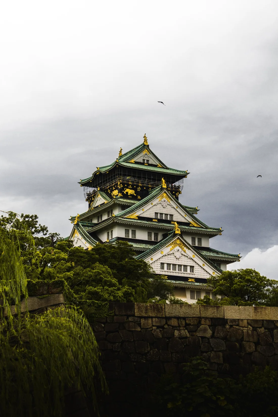 Tiered castle keep with gold-trimmed eaves rising above a stone wall and dark trees under a heavy grey sky.