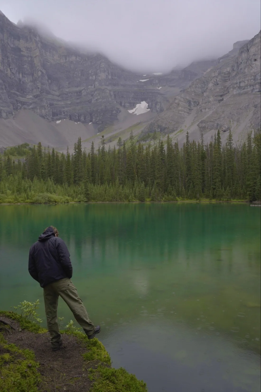 A person in a rain jacket standing on a mossy ledge above an emerald cirque lake, spruce forest and a cloud-covered rock wall beyond.