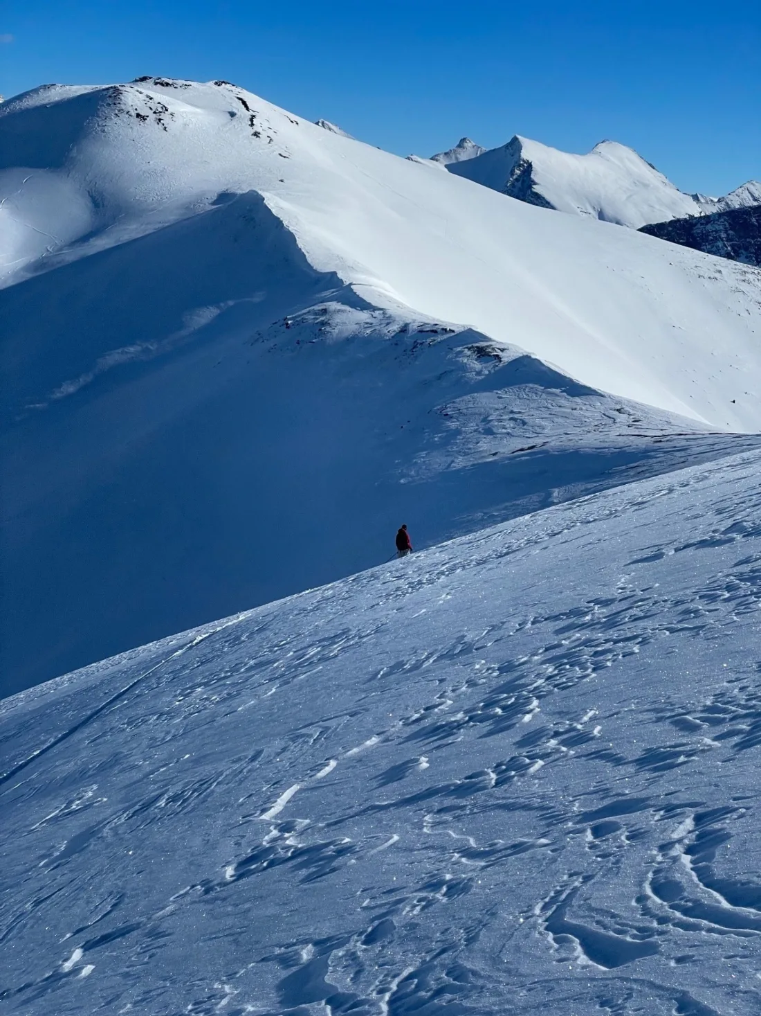 A single skier in a red jacket climbing a wind-scoured snow ridge, with corniced ridgelines and distant peaks under deep blue sky.