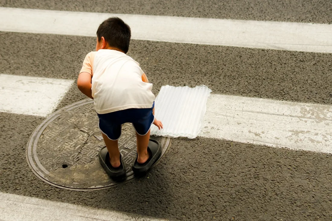 A child in a white shirt crouching on a painted crosswalk stripe beside a manhole cover, seen from behind.