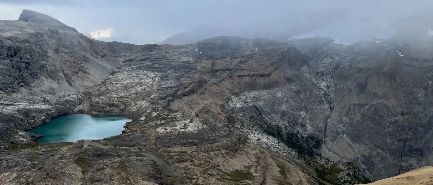 A lone hiker crossing a wide ochre scree slope high above a turquoise tarn, with grey limestone benches and a glacier lost in storm cloud beyond.