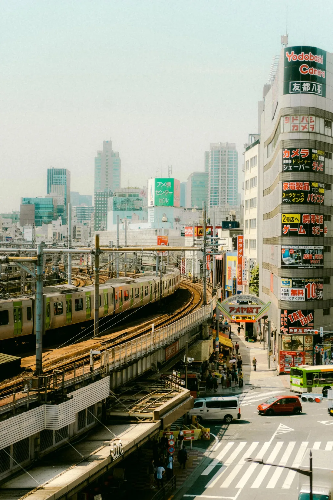 A commuter train on an elevated line curving between signboard-covered buildings above a busy city intersection.