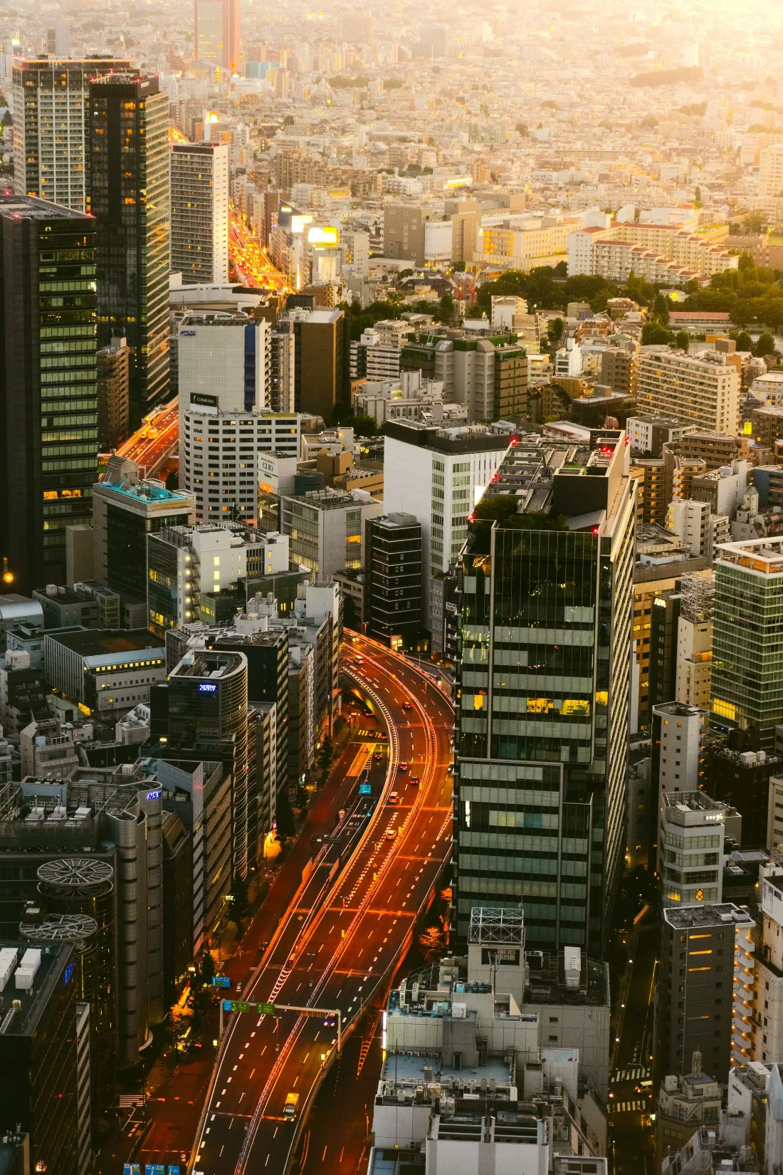 Aerial view over a dense city at dusk with an expressway cutting a lit orange line between towers to the horizon.