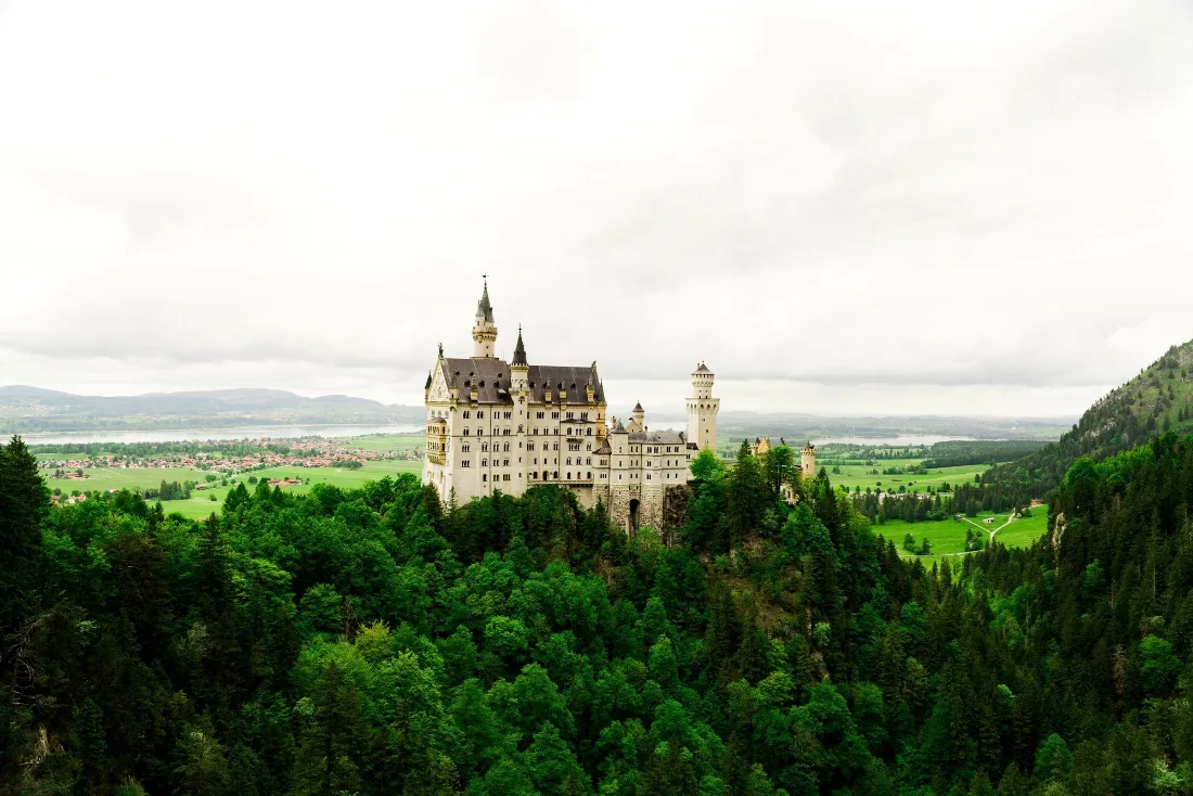A white turreted castle standing on a wooded hill above a wide green valley with pale sky behind.