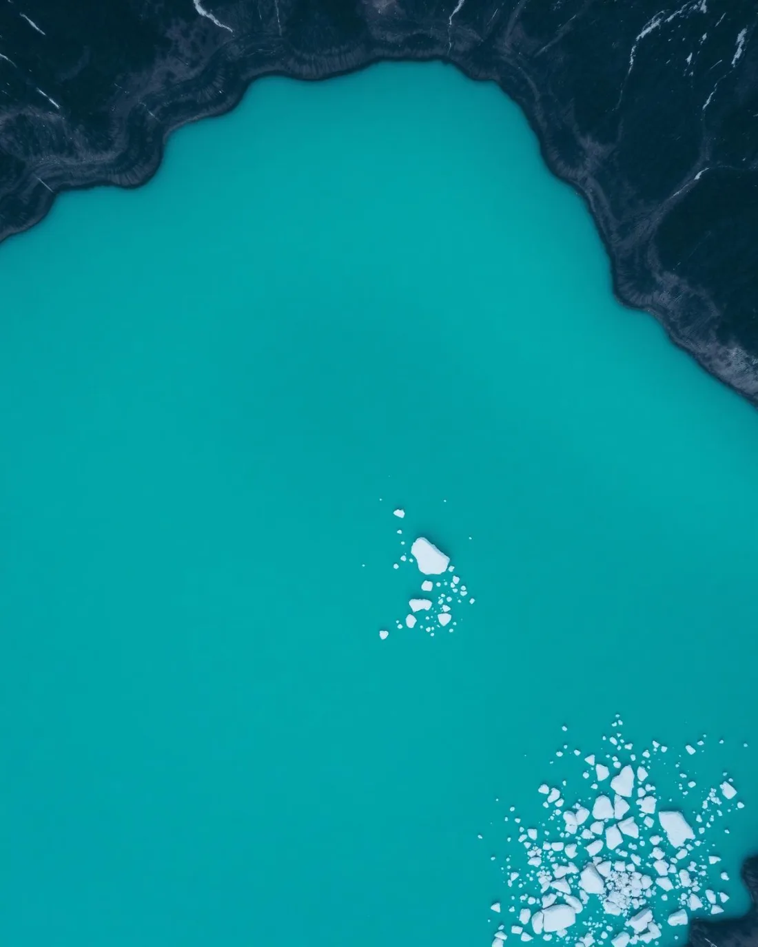 Aerial of a turquoise glacial lake carrying thin ice floes.
