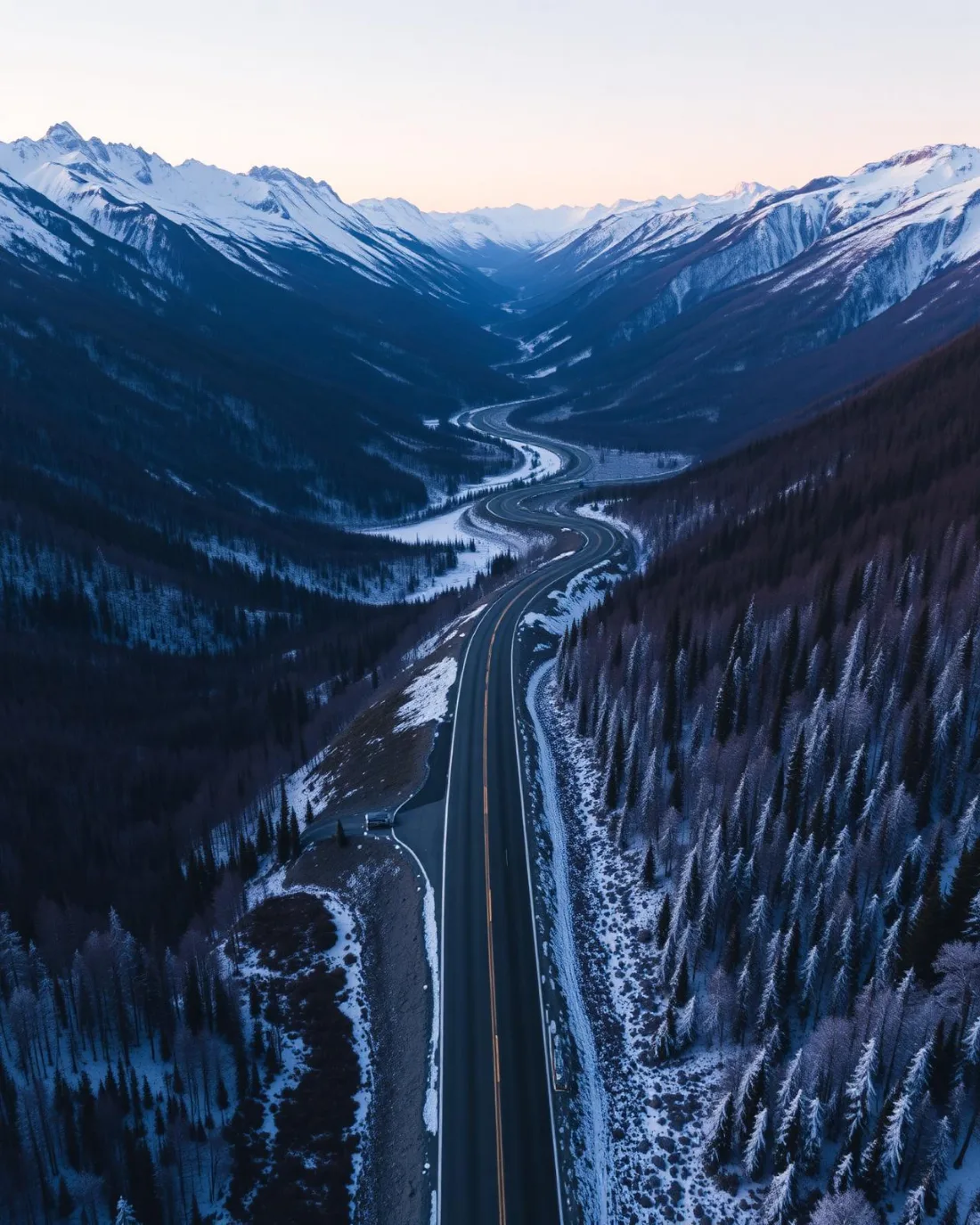 Aerial looking down a two-lane highway winding through a snow-dusted mountain valley at dusk, forested slopes falling away to snow-capped peaks.