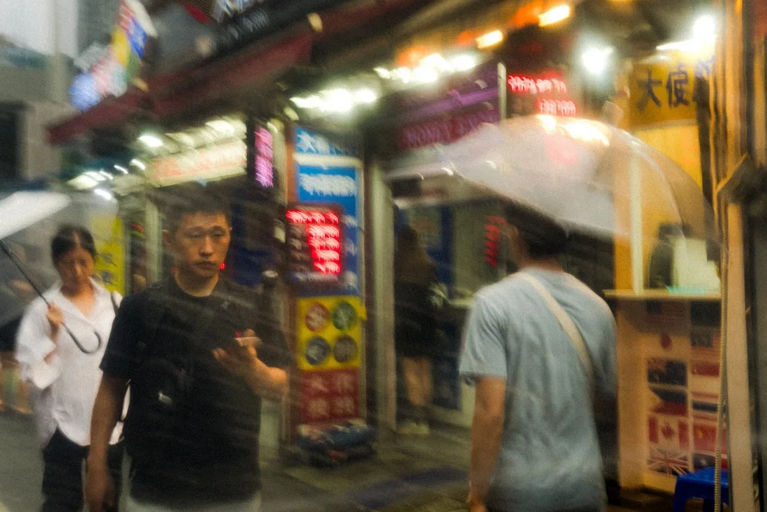 Passers-by crossing in front of a brightly lit street food stall at night, signage and awnings glowing above them.