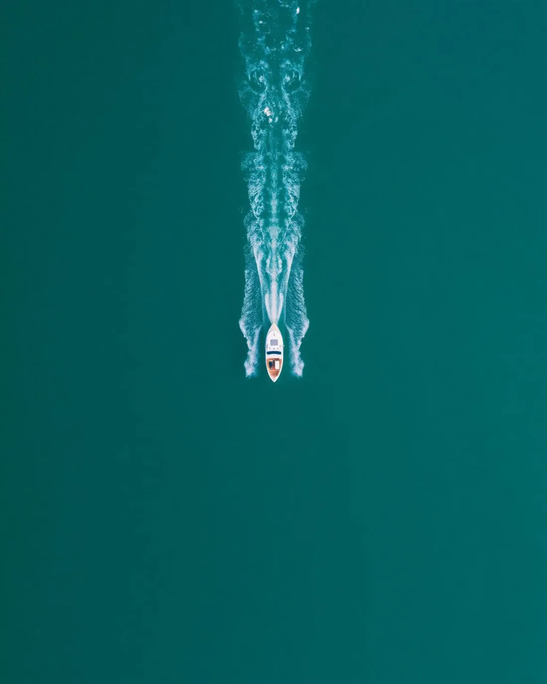 Single white boat cutting a wake straight across deep teal water, photographed from directly overhead.