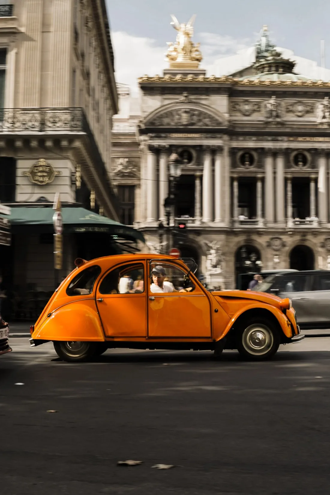 A bright orange vintage car parked on a plaza in front of an ornate stone façade with gilded sculpture above.