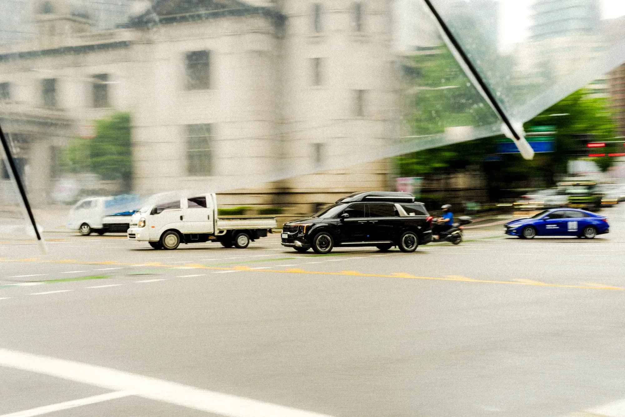 Panned exposure of vehicles crossing an intersection, the stone building behind smeared into horizontal streaks.