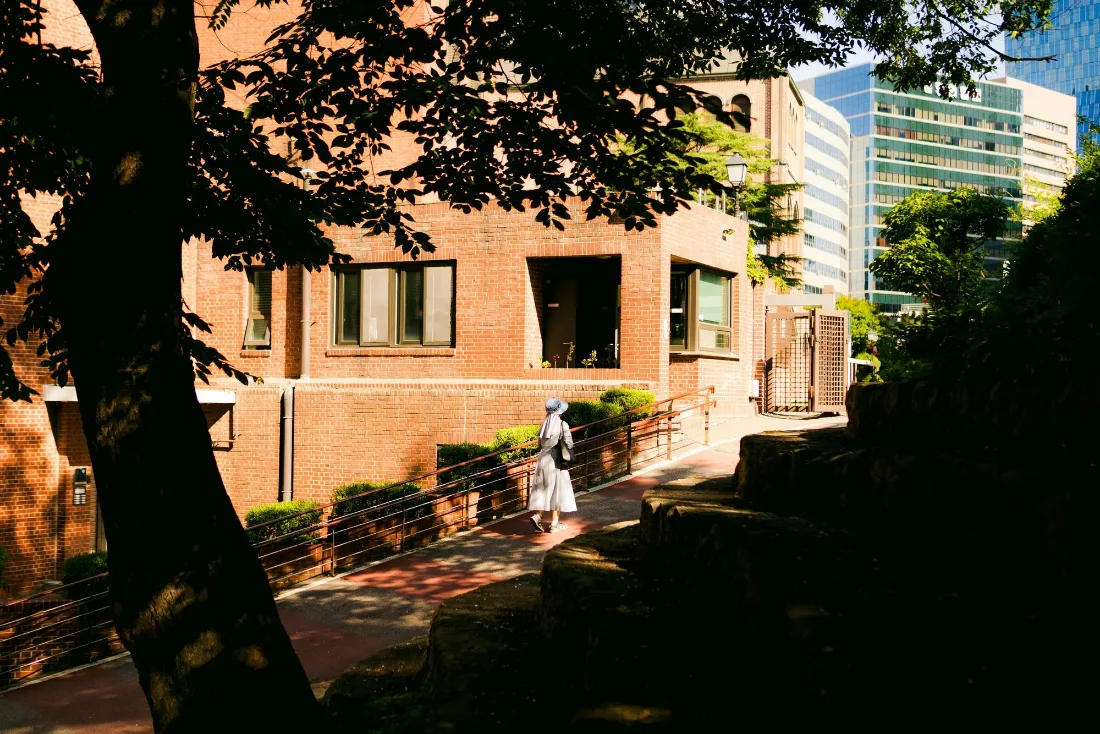 A single figure walking a shaded path beside an old brick building, glass towers catching sun in the gap beyond.