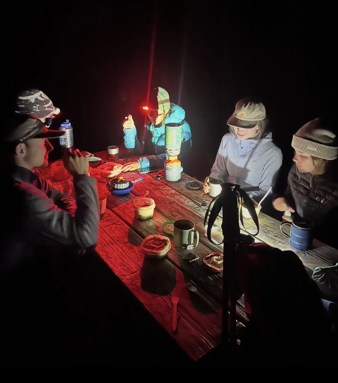 Four people eating around a wooden picnic table in the dark, lit only by red headlamps and the blue flame of a camp stove.