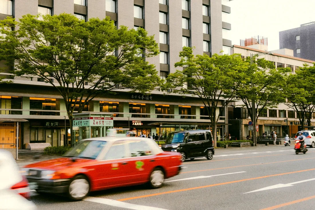 A red taxi pulling along a tree-lined boulevard past shopfronts and a tiled office block in flat midday light.