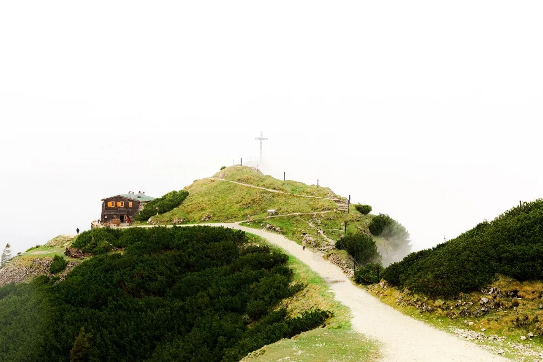 A dirt path climbing a grassy headland to a small building and a cross on the summit against a blown-out white sky.