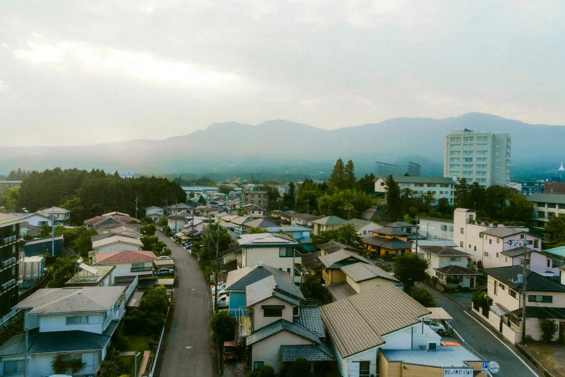 Metal and tile rooftops of a valley town stepping downhill, with rain cloud pressing on the mountains behind.