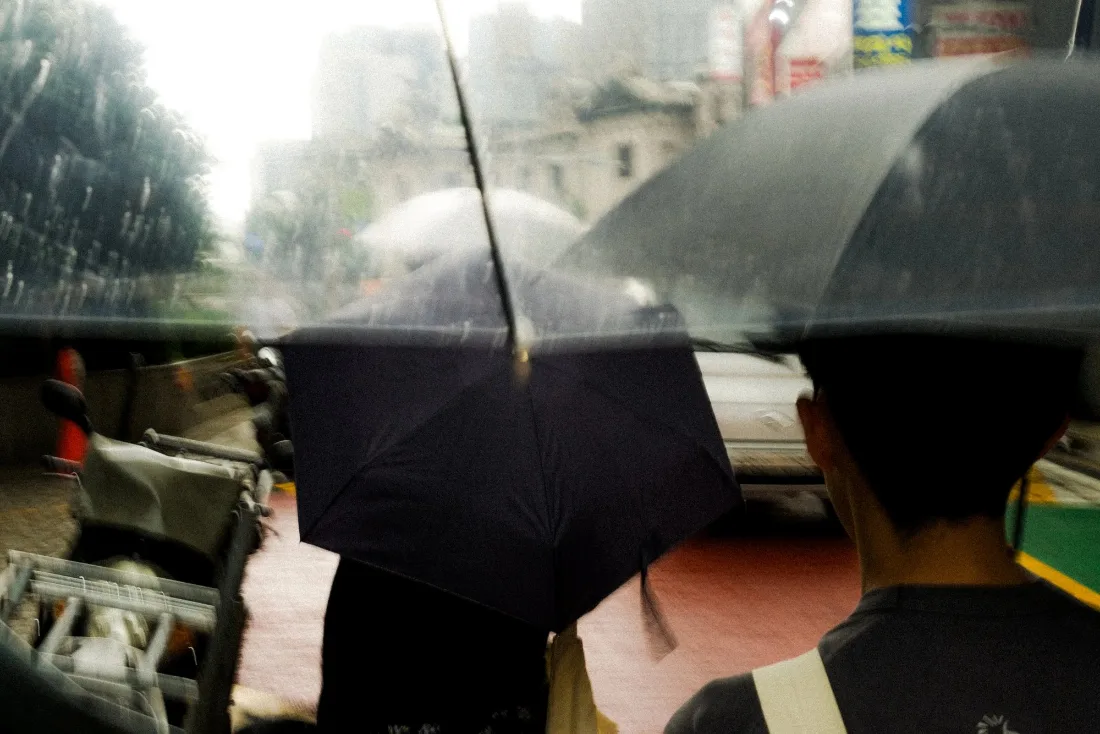 Clear and black umbrellas overlapping in the rain as people wait at a crowded city crossing, signage blurred behind.